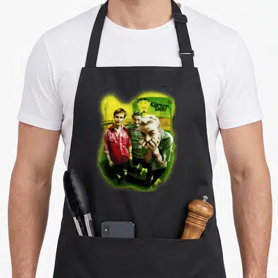 Funny neon novelty apron - Three men standing in front of Green Day text inside yellow organic shape — shown on a black cotton canvas apron against a white background