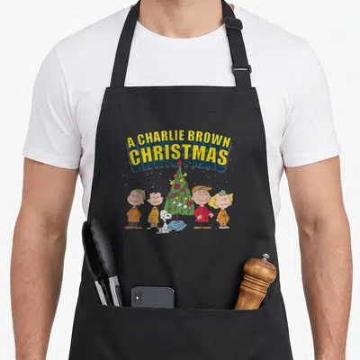 Charlie special aprons - Arched text above a Christmas tree surrounded by four children and a dog in cartoon style — shown on a black cotton canvas apron against a white background
