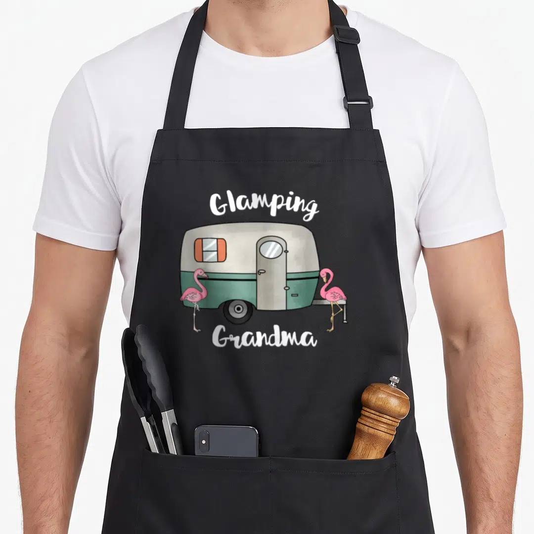 Glamping grandma apron - Vintage camper with two pink flamingos and Glamping Grandma text — shown on a black cotton canvas apron against a white background
