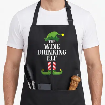 Drinking wine apron - Stacked text The Wine Drinking Elf with green hat and red striped legs — shown on a black cotton canvas apron against a white background