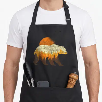 Outdoor bear apron - Retro bear silhouette facing right containing a forest and sun in brown, orange, yellow, and cream — shown on a black cotton canvas apron against a white background