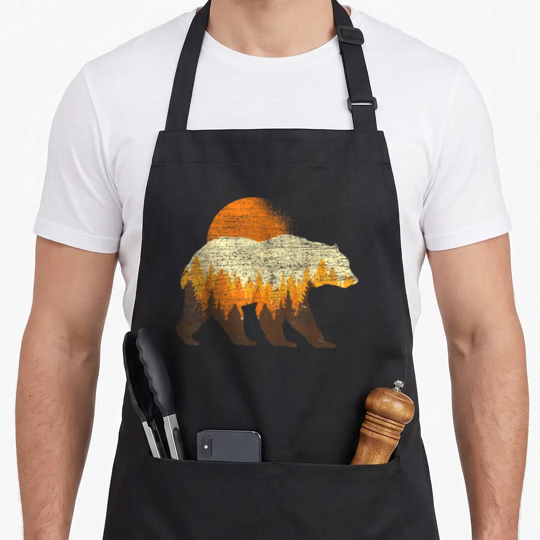 Outdoor bear apron - Retro bear silhouette facing right containing a forest and sun in brown, orange, yellow, and cream — shown on a black cotton canvas apron against a white background