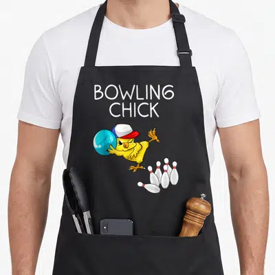 Chick bowling apron - Cartoon yellow chick wearing cap bowls blue ball at five white pins with red stripes — shown on a black cotton canvas apron against a white background