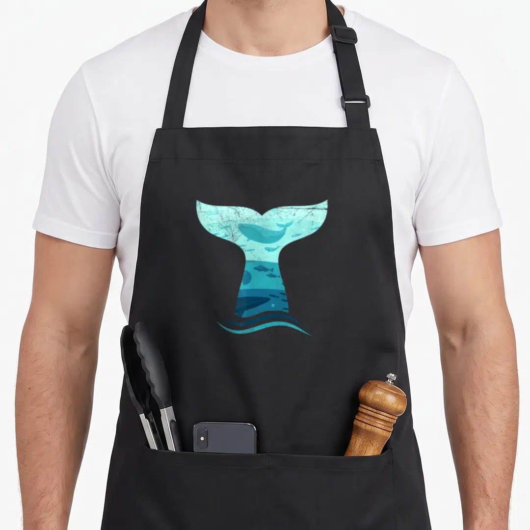 Whale orca apron - Large blue whale tail silhouette framing a swimming whale and fish scene with ocean waves below — shown on a black cotton canvas apron against a white background
