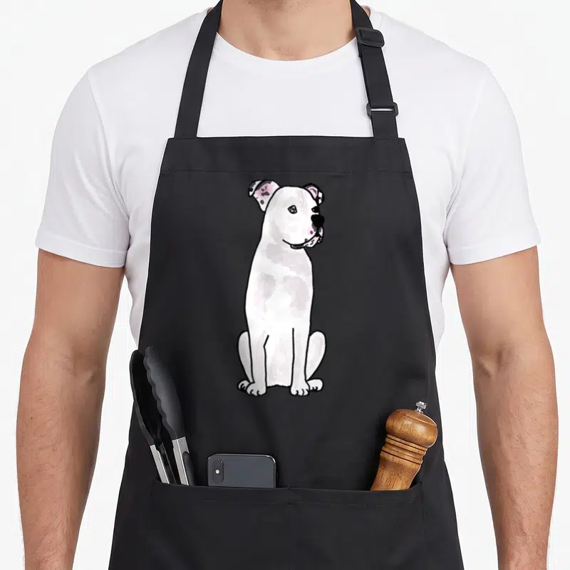 Bulldog smiletodaytees apron - Cartoon white dog sitting facing right with pink ears and black outlines — shown on a black cotton canvas apron against a white background