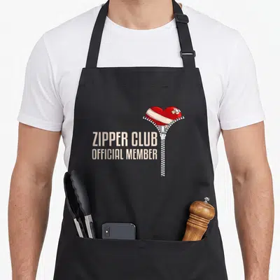 Surgery zipper open apron - Red heart with beige bandage and zipper next to stacked beige text reading ZIPPER CLUB — shown on a black cotton canvas apron against a white background