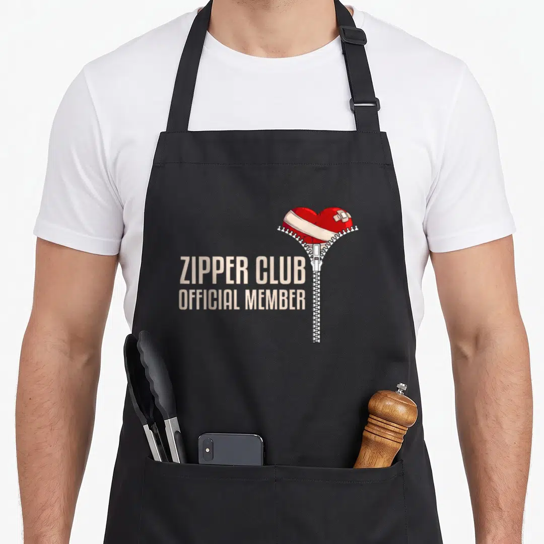 Surgery zipper open apron - Red heart with beige bandage and zipper next to stacked beige text reading ZIPPER CLUB — shown on a black cotton canvas apron against a white background