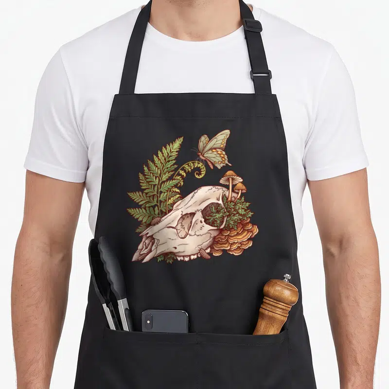 Deer witch apron - Cartoon skull with green fern, orange mushrooms, mossy eye socket, and crawling isopods — shown on a black cotton canvas apron against a white background