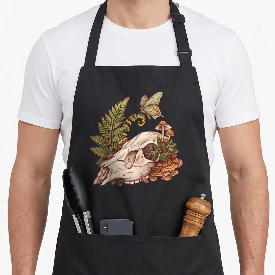 Deer witch apron - Cartoon skull with green fern, orange mushrooms, mossy eye socket, and crawling isopods — shown on a black cotton canvas apron against a white background