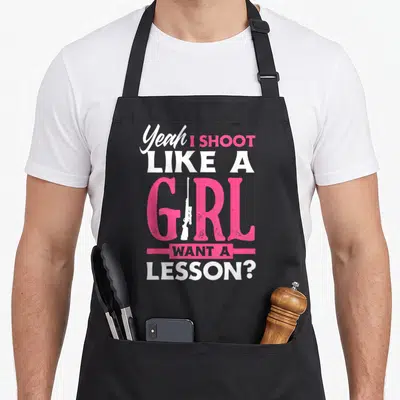 Yeah hunter apron - Pink and white stacked text reading Yeah I Shoot Like A Girl Want A Lesson with a rifle silhouette — shown on a black cotton canvas apron against a white background