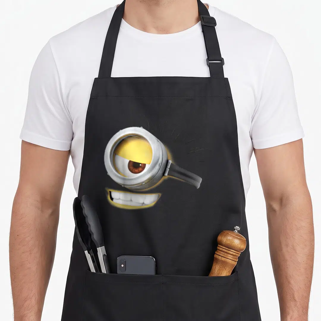 Carl face apron - Yellow minion eye with silver goggle and white teeth on black background — shown on a black cotton canvas apron against a white background
