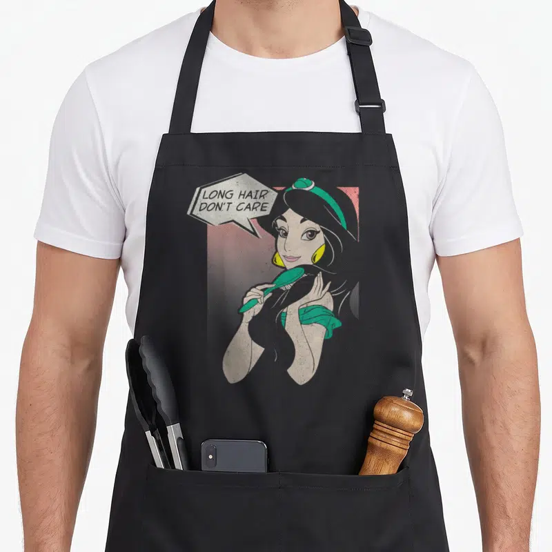 Jasmine hair apron - Cartoon Jasmine holding green brush with speech bubble text LONG HAIR DON'T CARE — shown on a black cotton canvas apron against a white background