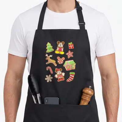 Gingerbread cooky women's christmas aprons - Mickey and Minnie gingerbread cookies with reindeer, house, and candy cane — shown on a black cotton canvas apron against a white background