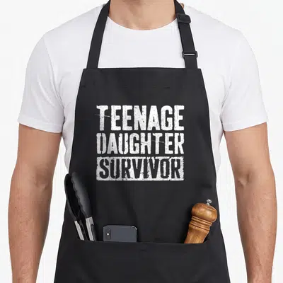 Teenage father day apron - White stacked text reading TEENAGE DAUGHTER SURVIVOR in distressed font — shown on a black cotton canvas apron against a white background