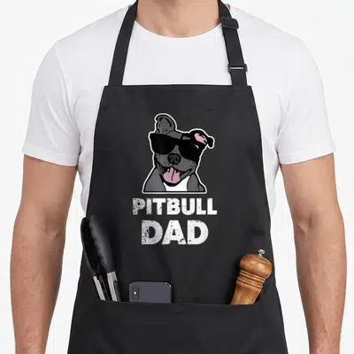 Bluenose dog dad apron - Cartoon grey pitbull with black sunglasses and pink tongue above white PITBULL DAD text — shown on a black cotton canvas apron against a white background