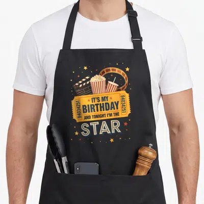Movie birthday family aprons - Retro yellow birthday ticket with star text surrounded by popcorn, drink, clapperboard — shown on a black cotton canvas apron against a white background