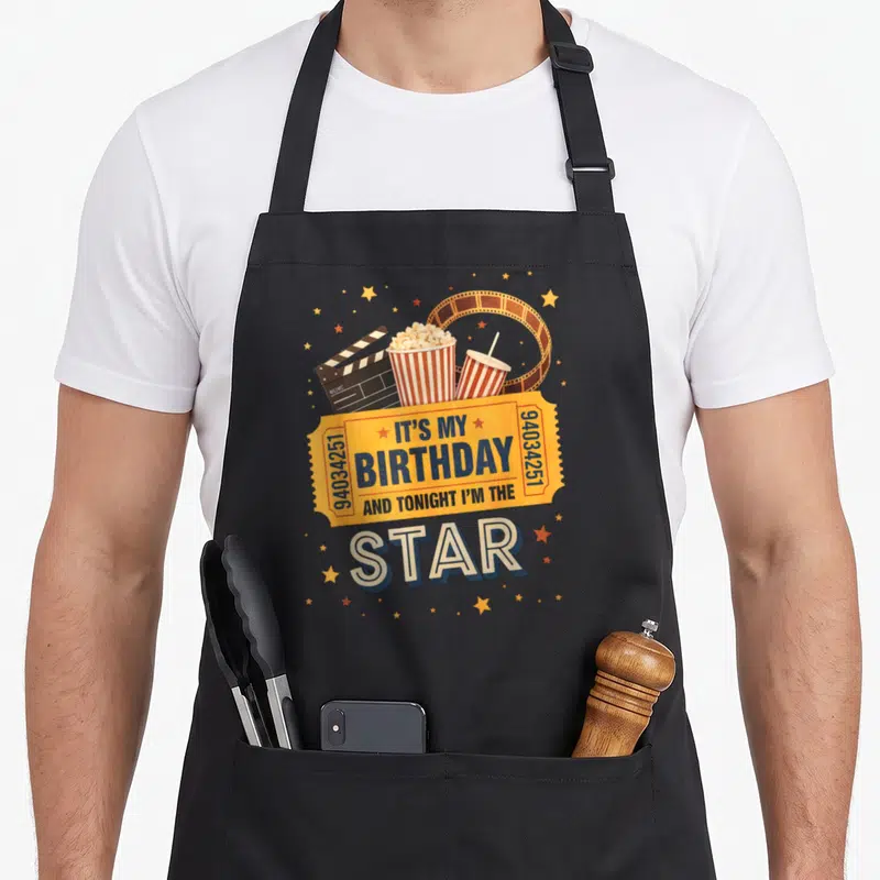 Movie birthday family aprons - Retro yellow birthday ticket with star text surrounded by popcorn, drink, clapperboard — shown on a black cotton canvas apron against a white background