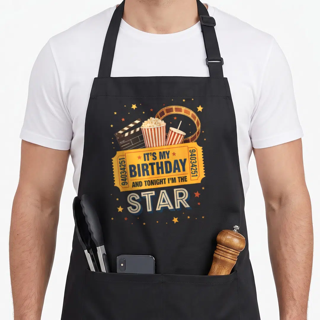 Movie birthday family aprons - Retro yellow birthday ticket with star text surrounded by popcorn, drink, clapperboard — shown on a black cotton canvas apron against a white background