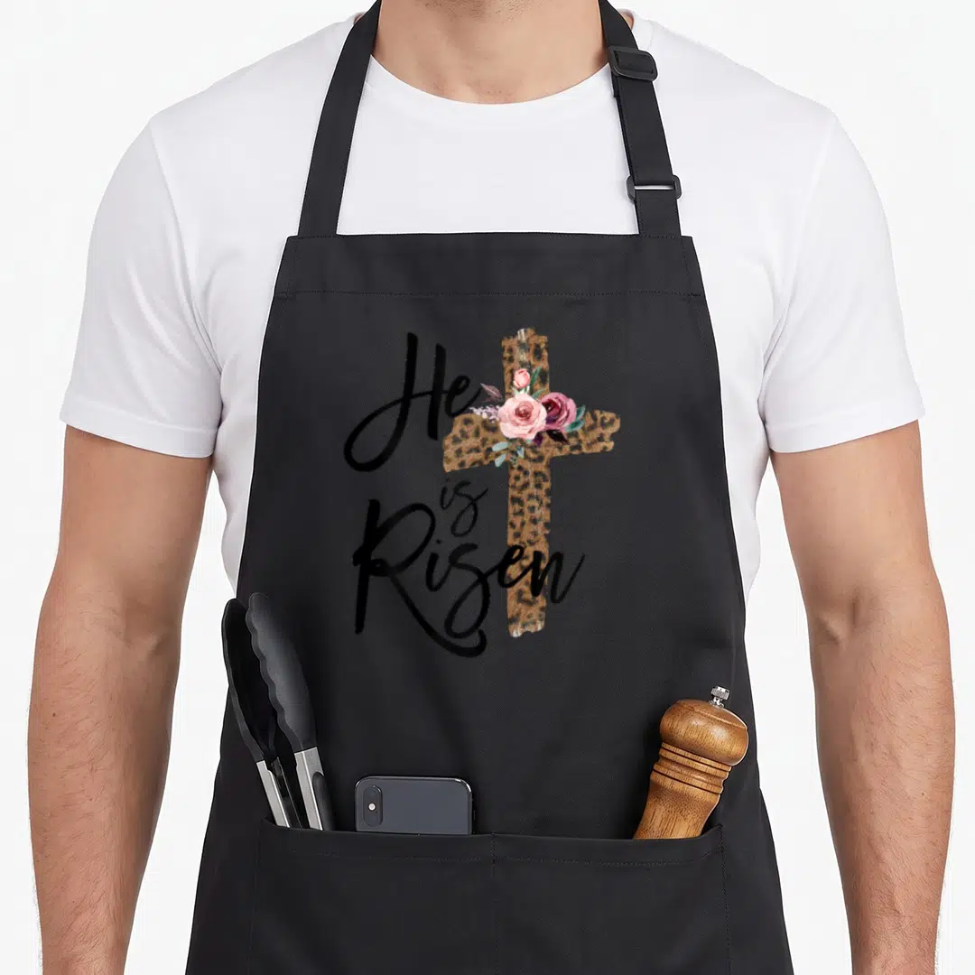Risen leopard apron - He is Risen text next to leopard cross with pink roses — shown on a black cotton canvas apron against a white background