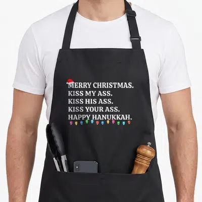 Christmas winter apron - Distressed white text reading Merry Christmas Kiss My Ass with Santa hat and multicolored — shown on a black cotton canvas apron against a white background