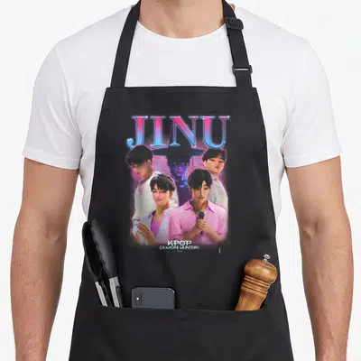 Jinu novelty apron - Four cartoon men and purple figure under large JINU text with KPOP DEMON HUNTERS label at bottom — shown on a black cotton canvas apron against a white background