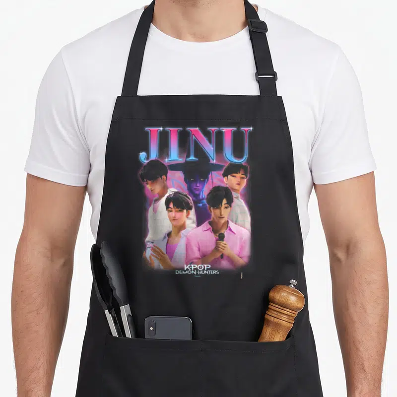 Jinu novelty apron - Four cartoon men and purple figure under large JINU text with KPOP DEMON HUNTERS label at bottom — shown on a black cotton canvas apron against a white background