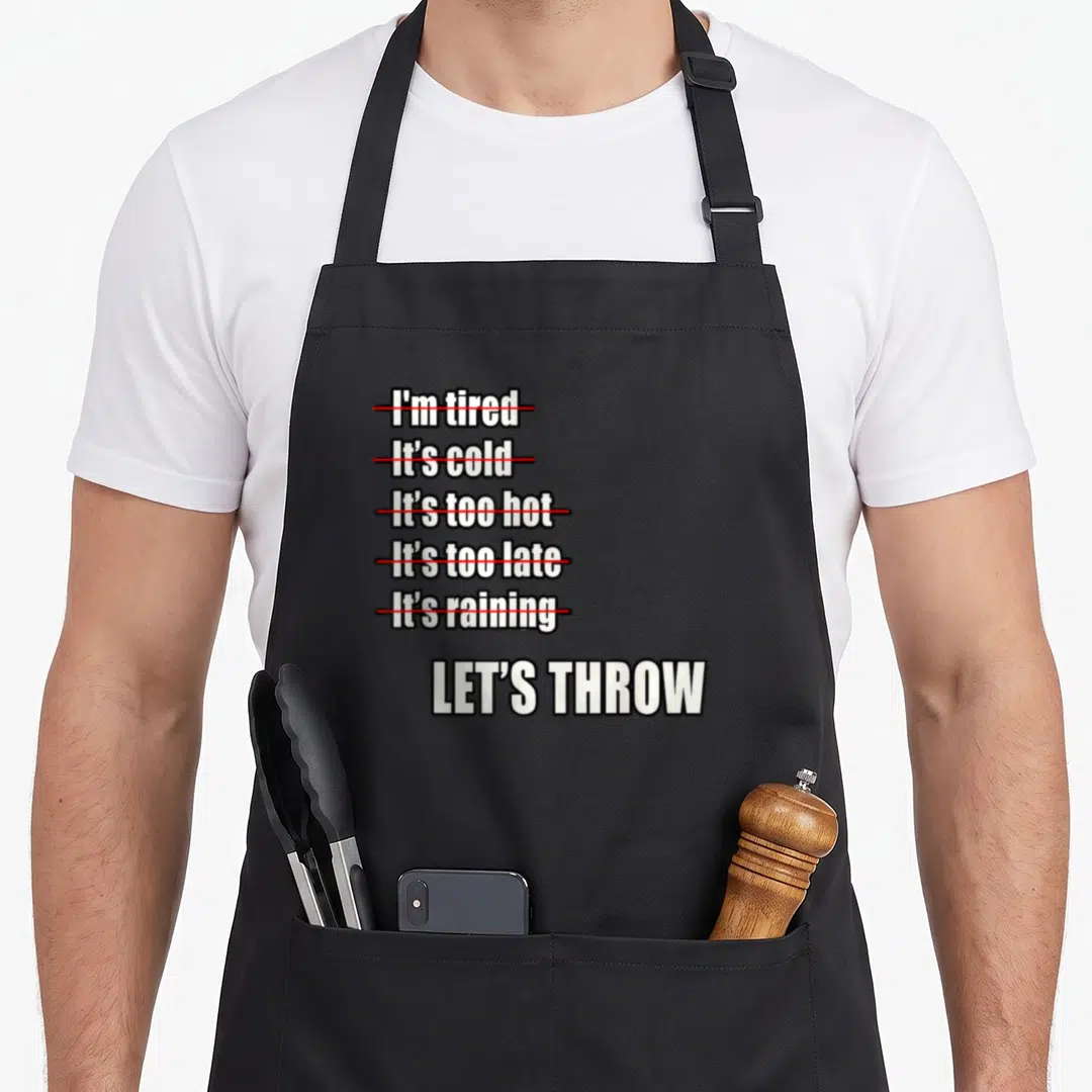Shot track apron - White text listing excuses crossed out in red above LET'S THROW slogan — shown on a black cotton canvas apron against a white background