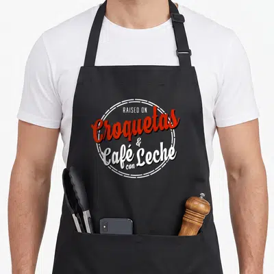 Raised croquetas apron - Red and white typography reading Raised on Croquetas and Cafe con Leche inside a dashed circle — shown on a black cotton canvas apron against a white background