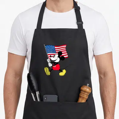 Americana flag apron - Cartoon Mickey Mouse walking right holding a waving US flag in red white and blue — shown on a black cotton canvas apron against a white background