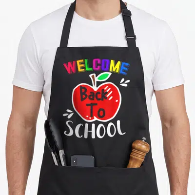 Welcome school teacher apron - Rainbow WELCOME Back To SCHOOL text with red apple and white sparkles — shown on a black cotton canvas apron against a white background