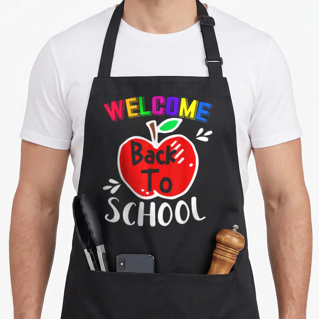 Welcome school teacher apron - Rainbow WELCOME Back To SCHOOL text with red apple and white sparkles — shown on a black cotton canvas apron against a white background