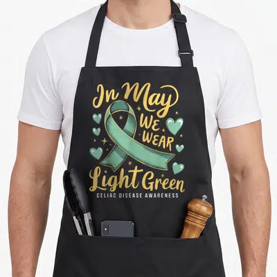 May light green apron - Green awareness ribbon surrounded by hearts and gold text reading In May We Wear Light Green — shown on a black cotton canvas apron against a white background