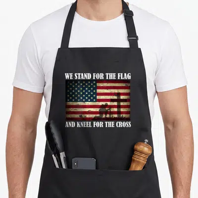 Stand cross back apron - Distressed USA flag with kneeling soldier silhouette and white text WE STAND FOR THE FLAG — shown on a black cotton canvas apron against a white background