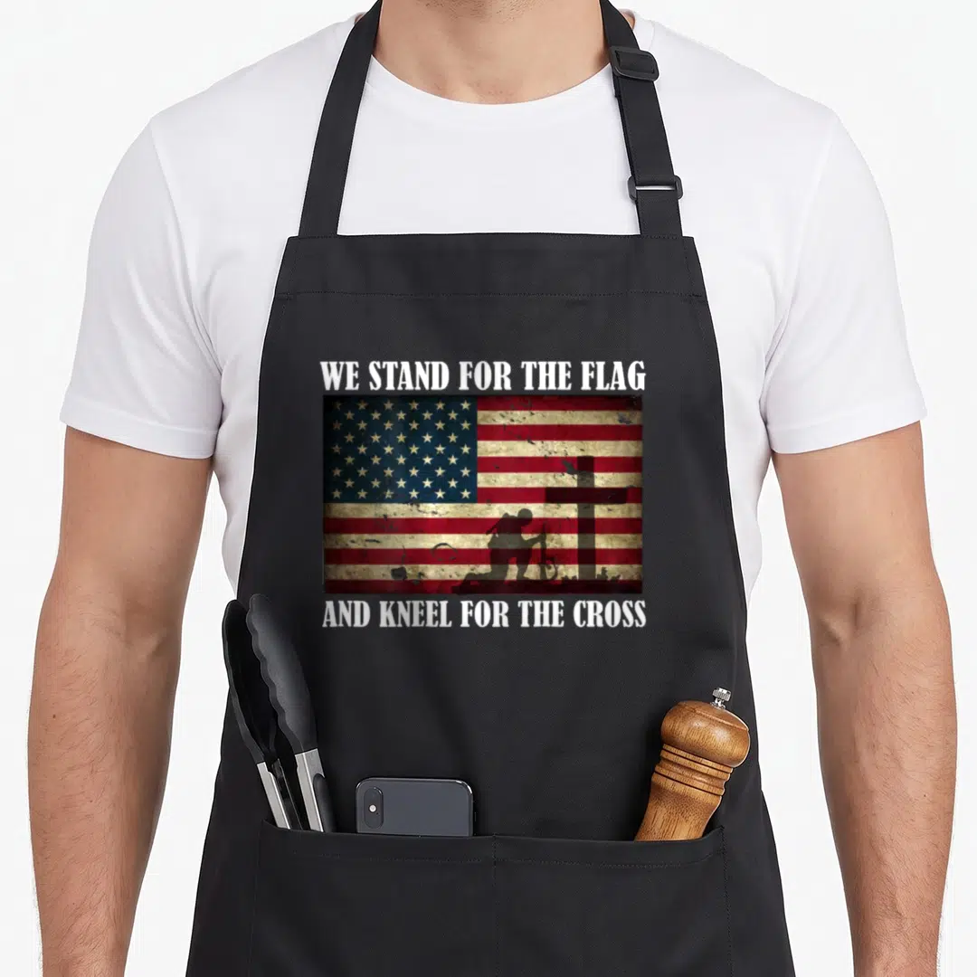 Stand cross back apron - Distressed USA flag with kneeling soldier silhouette and white text WE STAND FOR THE FLAG — shown on a black cotton canvas apron against a white background