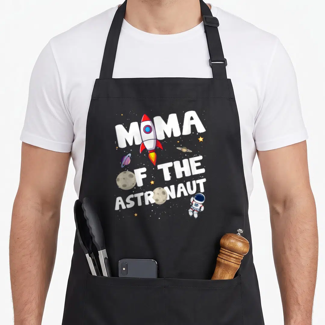 Space mama apron - MOMA OF THE ASTRONAUT text with rocket inside letters and scattered planets — shown on a black cotton canvas apron against a white background
