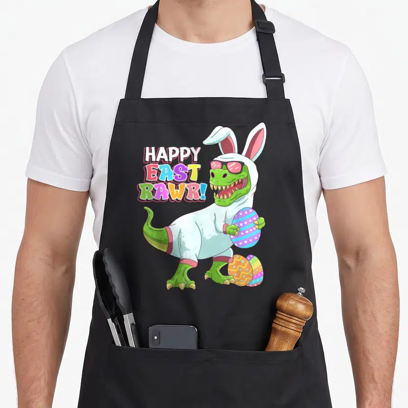 Eastrawr easter apron - Dinosaur in bunny costume holding striped egg with HAPPY EAST RAWR text — shown on a black cotton canvas apron against a white background