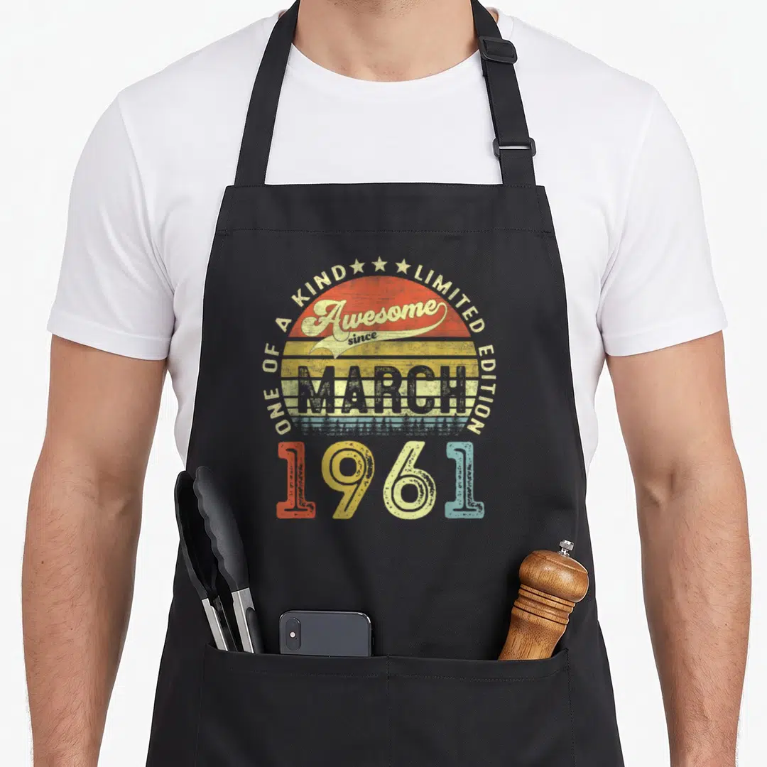 March 60th birthday apron - Retro sunburst with Awesome since MARCH 1961 text in red yellow blue orange stripes — shown on a black cotton canvas apron against a white background