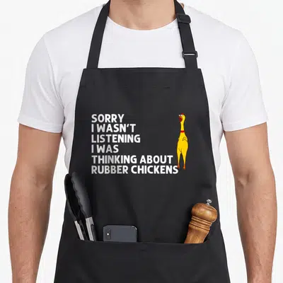 Squeaking chicken apron - White text stacking beside a yellow cartoon chicken on a black bib — shown on a black cotton canvas apron against a white background