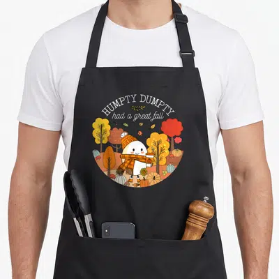 Leave cute fall aprons - Humpty Dumpty cartoon holding coffee in orange and yellow autumn scene with pumpkins — shown on a black cotton canvas apron against a white background