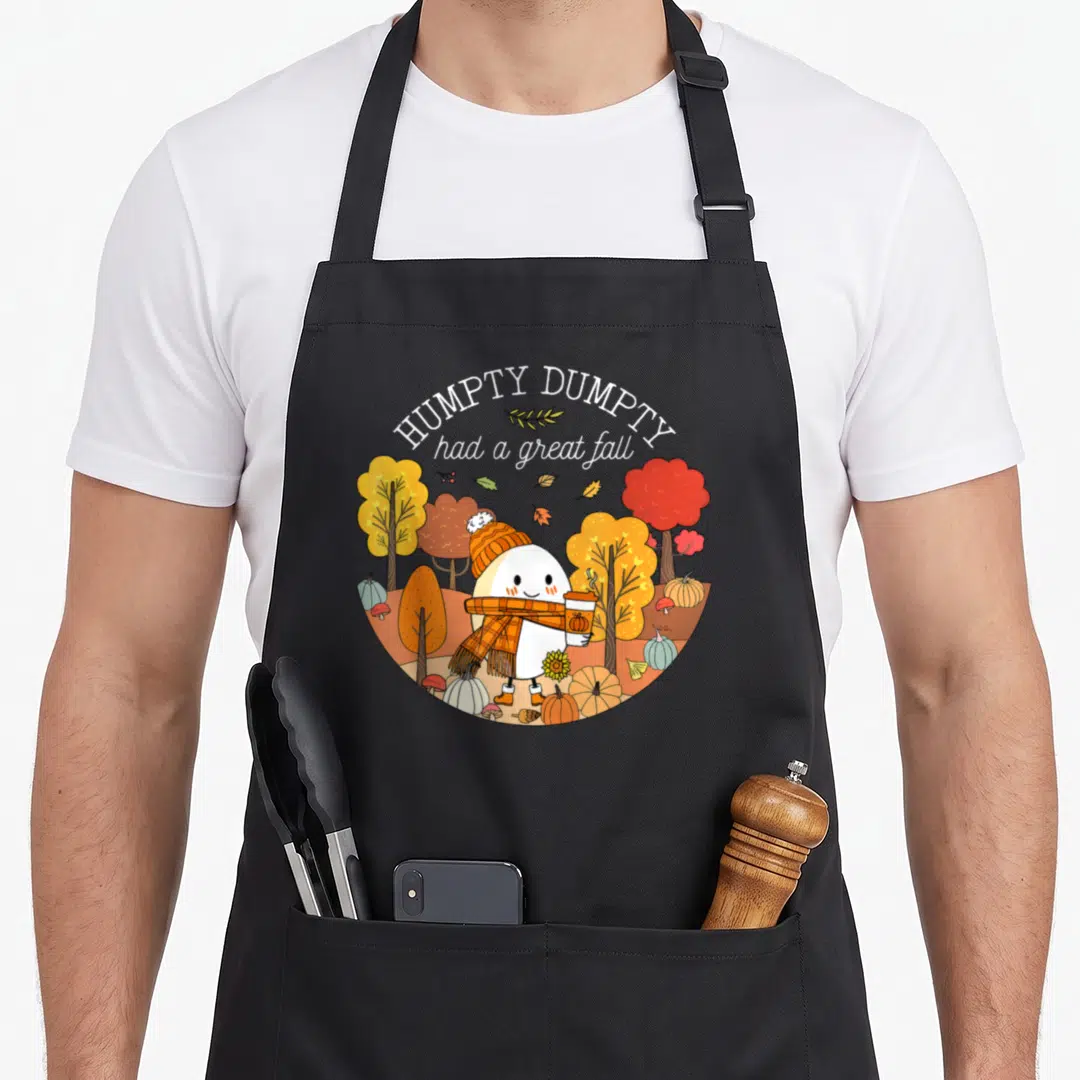 Leave cute fall aprons - Humpty Dumpty cartoon holding coffee in orange and yellow autumn scene with pumpkins — shown on a black cotton canvas apron against a white background