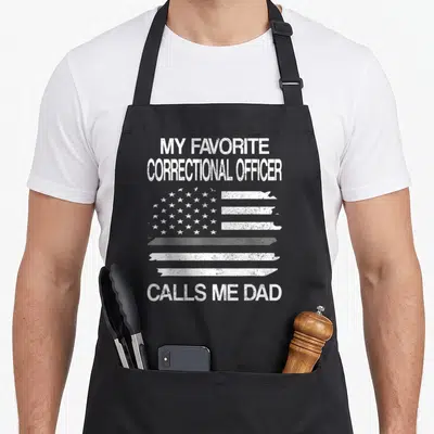 Correctional officer dad apron - White distressed US flag graphic with text MY FAVORITE CORRECTIONAL OFFICER CALLS ME — shown on a black cotton canvas apron against a white background