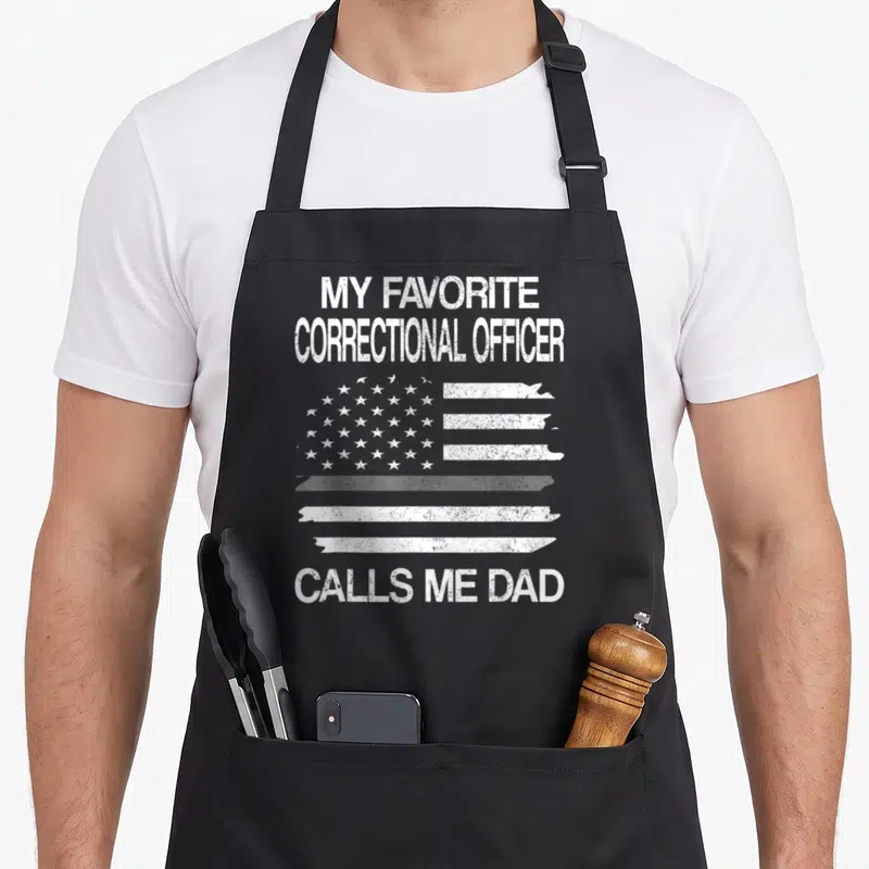 Correctional officer dad apron - White distressed US flag graphic with text MY FAVORITE CORRECTIONAL OFFICER CALLS ME — shown on a black cotton canvas apron against a white background