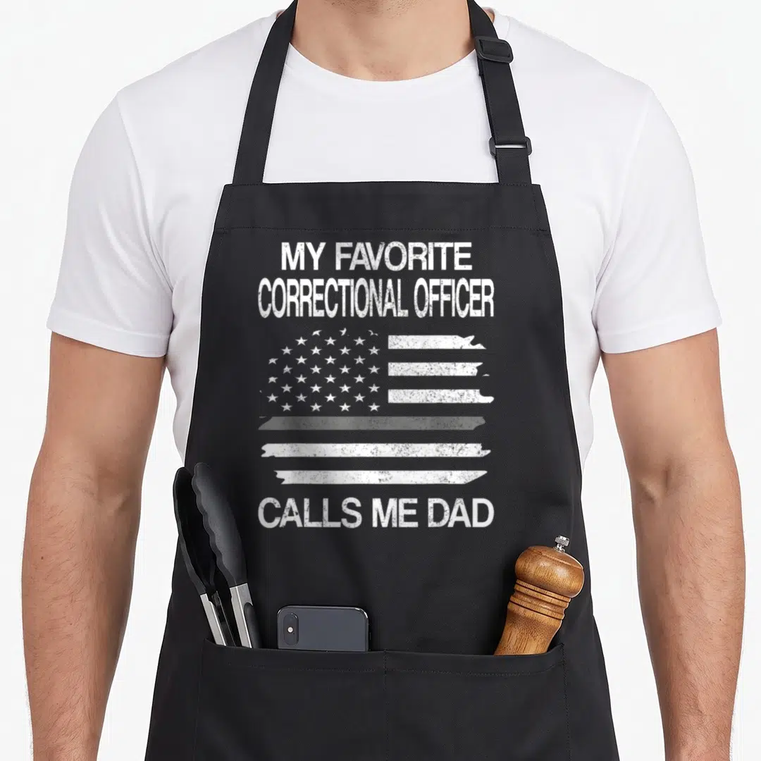 Correctional officer dad apron - White distressed US flag graphic with text MY FAVORITE CORRECTIONAL OFFICER CALLS ME — shown on a black cotton canvas apron against a white background