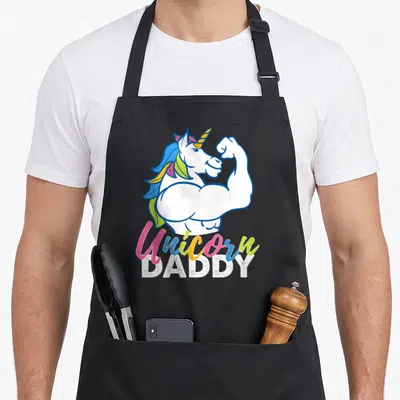 Magical daddy apron - Cartoon muscular unicorn flexing with colorful Unicorn DADDY text — shown on a black cotton canvas apron against a white background