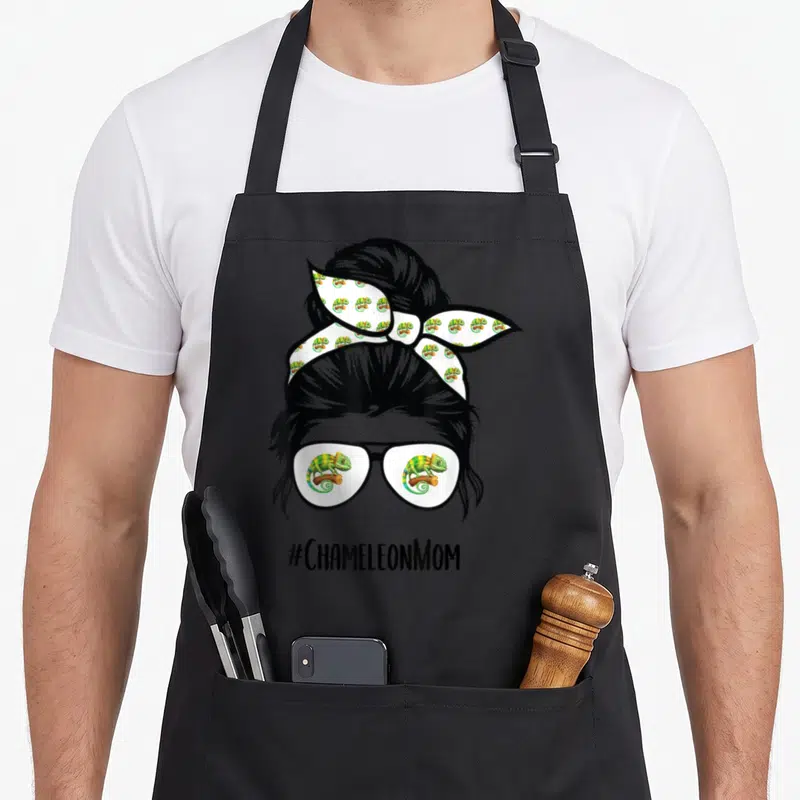 Chameleon messy aprons - Woman silhouette with chameleon glasses bun and #CHAMELEONMOM text — shown on a black cotton canvas apron against a white background