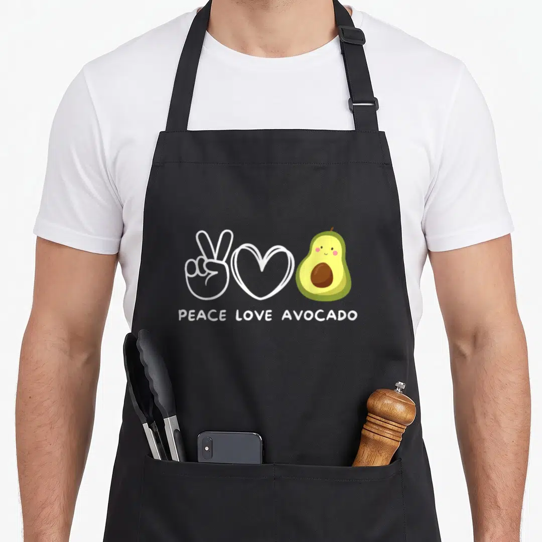 Peace avocado apron - White peace sign, heart, and cartoon avocado with text below — shown on a black cotton canvas apron against a white background