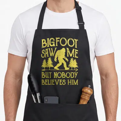 But bigfoot apron - Yellow distressed text and Bigfoot silhouette walking between pine trees with arrow — shown on a black cotton canvas apron against a white background