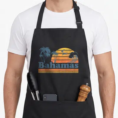 Bahamas 70s apron - Retro beach scene with striped sun, blue palms, birds, wave, and Bahamas text — shown on a black cotton canvas apron against a white background