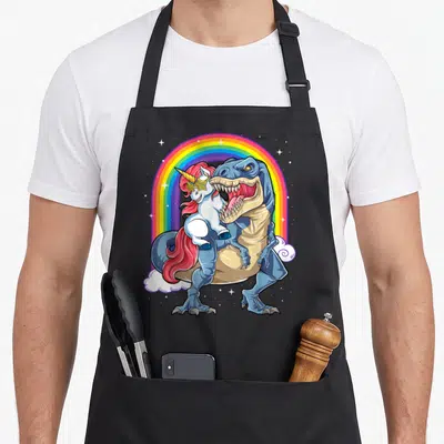 Rex rainbow dinosaur apron - Blue T-Rex carrying pink-maned unicorn under rainbow arch with stars and clouds — shown on a black cotton canvas apron against a white background