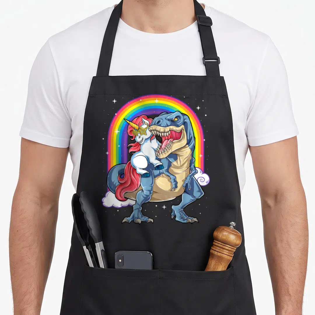 Rex rainbow dinosaur apron - Blue T-Rex carrying pink-maned unicorn under rainbow arch with stars and clouds — shown on a black cotton canvas apron against a white background